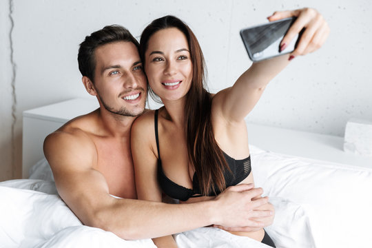 Image of caucasian couple man and woman taking selfie photo together, while lying in bed at home or hotel apartment