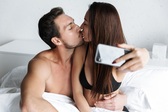 Image of attractive couple man and woman taking selfie photo together, while lying in bed at home or hotel apartment