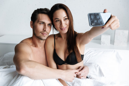 Image of european couple man and woman taking selfie photo together, while lying in bed at home or hotel apartment