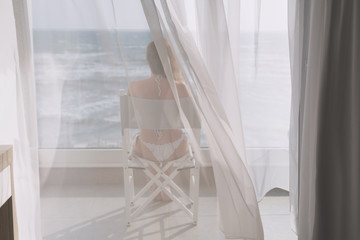 Naked beautiful female in the lace bikini sitting on the chair behind the transparent curtain on the glass balcony against the backdrop of the sea
