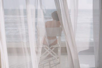 Naked beautiful female in the lace bikini sitting on the chair behind the transparent curtain on the glass balcony against the backdrop of the sea