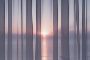 Window of a balcony with transparent curtains and view of morning sun and sea