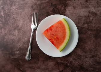 Slice of watermelon on a white plate with a fork to the side atop a maroon table top.