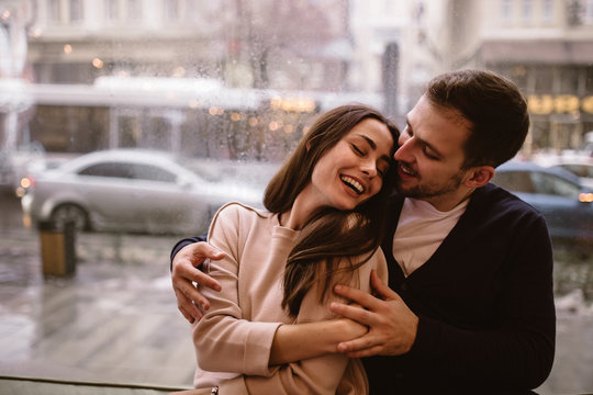 Loving guy hugs his beautiful happy girlfriend sitting on the windowsill in a cozy cafe.