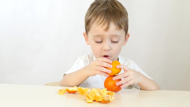 Cute little boy sitting at the table on a white background. The child picks up two ripe mandarins and puts it over his eyes, playing and laughing.