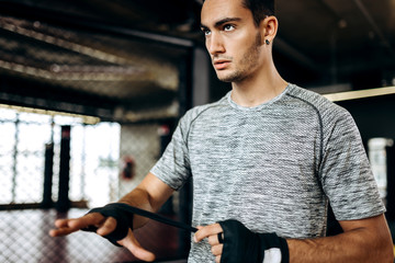 Dark-haired guy dressed in the grey t-shirt and black shorts stands  in the gym next the boxing ring and wraps a hand bandage on his hand