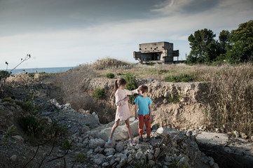 thin little girl refugee soothe her small brother on ruins of demolished building in war zone