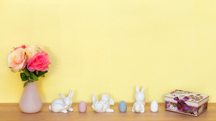 Eine Blumenvase mit Rosen und drei weiße Hasen mit drei Ostereier und einem Geschenk auf einem Holzboden