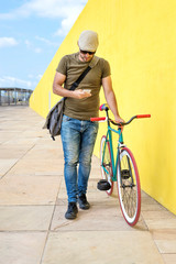 Fototapeta na wymiar Front view of a young trendy man with a fixed bike wearing casual clothes while using a mobile phone against a yellow wall outdoors in a sunny day