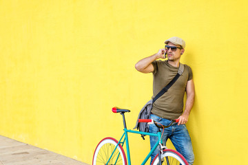 Naklejka premium Side view of a young adult man with a vintage bike and wearing casual clothes and sunglasses standing against a yellow wall while using a mobile phone in a sunny day