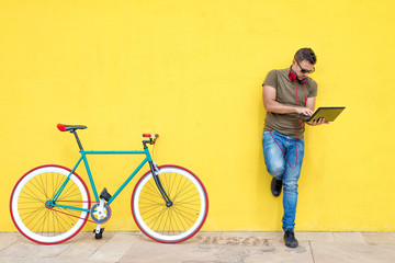Front view of a young trendy man with a fixed bike wearing casual clothes while using a laptop standing against a yellow wall outdoors in sunny day