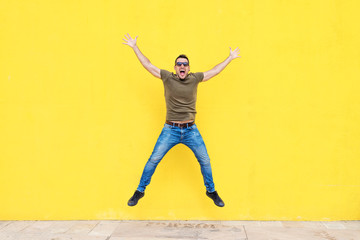 Fototapeta na wymiar Front view of a young man wearing sunglasses jumping against a yellow bright wall in a sunny day