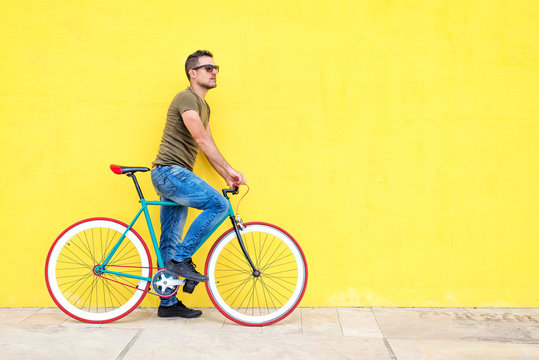 Side view of a young hipster man with a fixed bike wearing casual clothes while looking away against a yellow wall outdoors in a sunny day