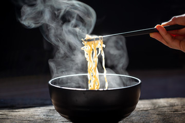 Chopsticks to tasty noodles with steam and smoke in bowl on wooden background, selective focus.,...