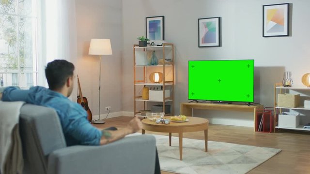 In the Living Room: Guy Relaxing on a Couch Watching Green Chroma Key Screen Television. 