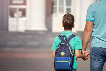 Father leads a little child school boy go hand in hand. Parent and son with backpack behind the...