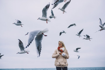 Young beautiful woman feeds seagulls on the sea. Pretty female wearing coat, scarf and watching...