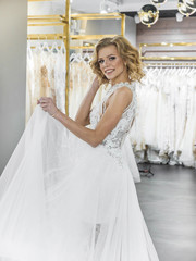 Naklejka premium Woman choosing wedding dress in shop. Attractive young bride is smiling while choosing wedding dress in modern wedding salon. Future wife. Woman surrounded by wedding dresses.