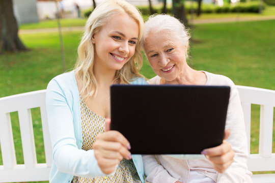 family, technology and people concept - happy smiling young daughter with tablet pc computer and senior mother sitting on park bench