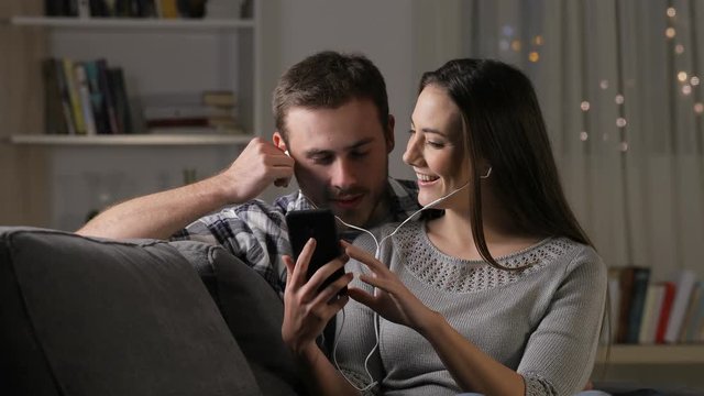 Happy couple listening to music sharing earphones sitting on a couch at home in the night
