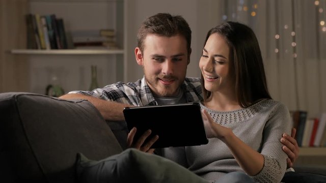 Happy couple watching online streaming videos on a tablet sitting on a couch in the night