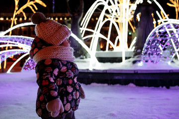 a cute little girl standing on a city square, leaning against Christmas light decorations during a night walk through the city. The girl enjoys illuminated Christmas decorations.