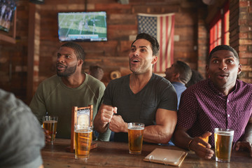 Group Of Male Friends Celebrating Whilst Watching Game On Screen In Sports Bar