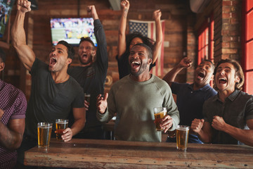 Group Of Male Friends Celebrating Whilst Watching Game On Screen In Sports Bar - obrazy, fototapety, plakaty
