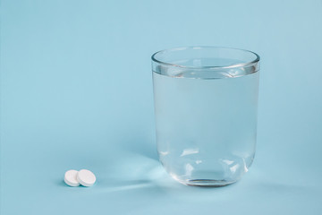 Transparent glass of drinking water and two white pills on a blue background. To get sick, take pills or undergo treatment. Concept of health care and pharmaceuticals. Front view and copy space