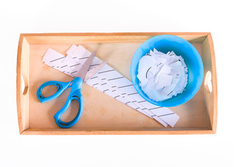Children's hands hold a wooden tray with montessori materials for a lesson from the practical life zone. Blue scissors, bowl, sheets for cutting. Isolate on white background.