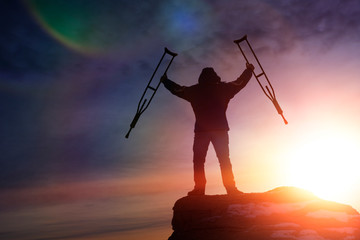 man holding crutches on top of the mountain against the evening sky. climbing to the top. achieving the goal, overcoming.