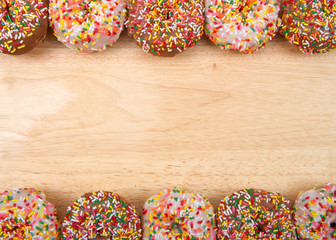 Flat lay view of many frosted donuts, chocolate and vanilla with candy sprinkles arranged in a border top and bottom on a light wood table with copy space.