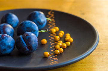 still life - juicy blue plums and sea buckthorn berries in a ceramic plate on a wooden background