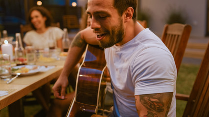 Big Family Garden Party Celebration, Gathered Together at the Table Relatives and Friends, Young and Elderly are Eating, Drinking, Passing Dishes, Joking and Having Fun. Decorative Lights Turned on.