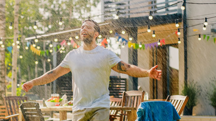 Handsome Fit Young Man Enjoys Summer Under the Rain Made by Garden Water Hose Sprinkler, Raises...