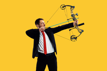 Businessman aiming at target with bow and arrow, isolated on yellow studio background. The...