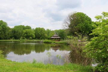 Obraz premium Midwest nature background with city park view. Beautiful late spring cloudscape with trees around the pond and wooden gazebo in a city park. Lakeview park, Middleton, Madison area, WI, USA.