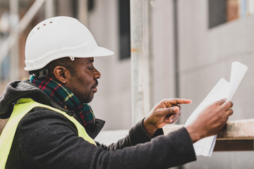 Absorbed African American male engineer wearing safety jacket and helmet checking technical...