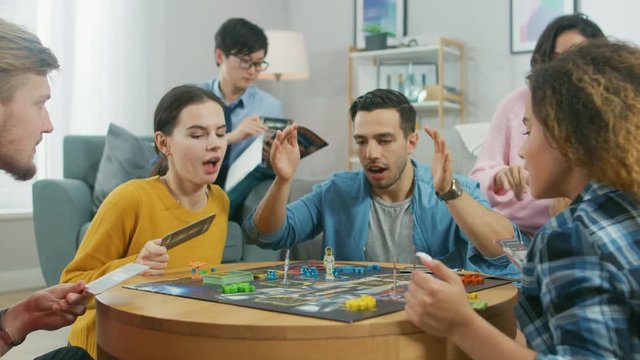 Diverse Group of Guys and Girls Playing in a Strategic Board Game with Cards and Dice. Man Throws Dice Impressively. Cozy Living Room in a Daytime
