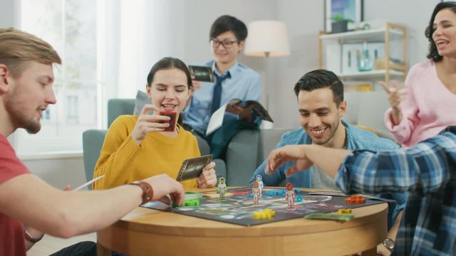 Diverse Group of Guys and Girls Playing in a Strategic Board Game with Cards and Dice. Man Throws Dice Impressively. Cozy Living Room in a Daytime