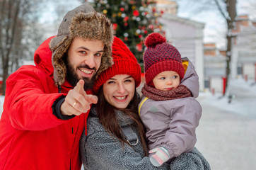 Obraz premium young energetic family. Husband and wife hug their child on the street in the middle of a snow-covered winter in a park near a tree, dressed in things of red color.