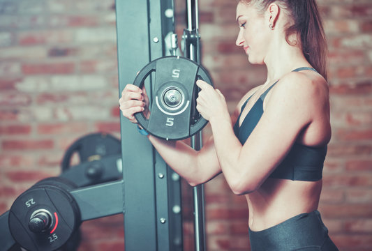 Fitness woman putting weight disk to barbell  against brick wall in gym