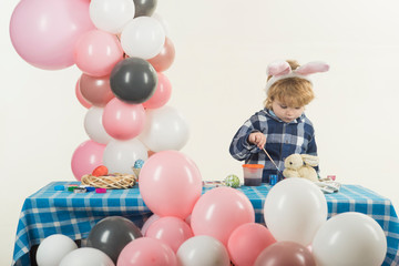 Fototapeta na wymiar Cute little boy plays with paints and washes brush in water. Little boy wears pink bunny ears. Pink, grey and white balloons on background. Easter eggs, straw bowl and tiny rabbit are on table