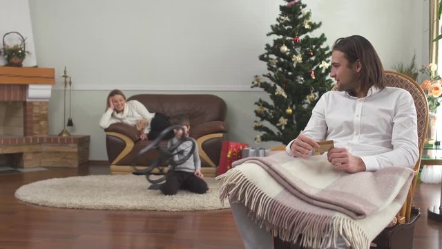 Handsome man sitting in arm-chair holding credit card. Mother and little son in the background. Happy family celebrates xmas at home. Christmas shopping concept.