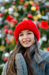 young, pretty model brunette poses for a photo in the winter near a Christmas tree in the middle of a winter city park, dressed in a red knitted hat and a warm gray coat and gloves