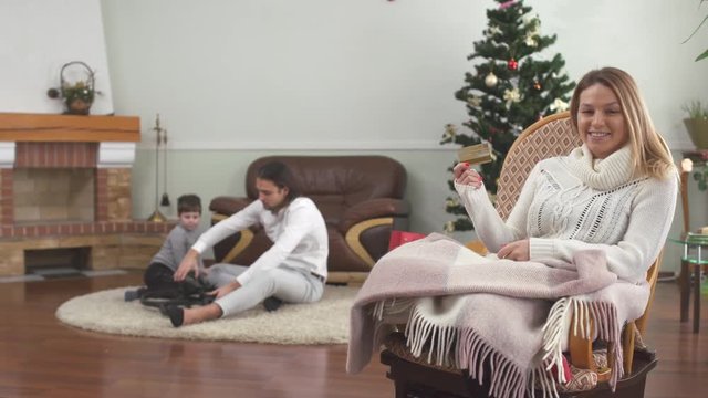 Pretty woman sitting in rocking chair holding credit card. Father and little son are playing with drone. Happy family celebrates xmas at home. Christmas shopping concept.