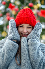 young, pretty model brunette poses for a photo in the winter near a Christmas tree in the middle of a winter city park, dressed in a red knitted hat and a warm gray coat and gloves