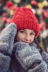 young, pretty model brunette poses for a photo in the winter near a Christmas tree in the middle of a winter city park, dressed in a red knitted hat and a warm gray coat and gloves