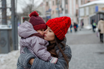 mother hugs and kisses her little baby, son or daughter, holding her in the middle of a snowy day in the city. A modern young mother wears a baby in her arms