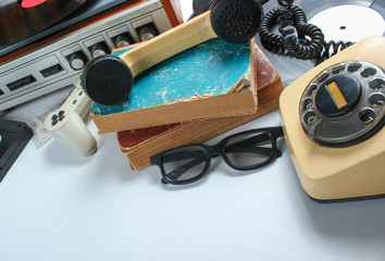 Retro 80s pop culture media objects on white background. Copy space. Sneakers, rotary phone, vinyl player, old books, audio, video tapes, 3d glasses, gamepad.